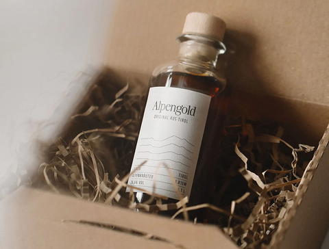 Alpengold Heuschnaps-Flasche mit hellem Etikett liegt in Geschenkbox mit Holzwolle, Kork und bernsteinfarbenem Inhalt