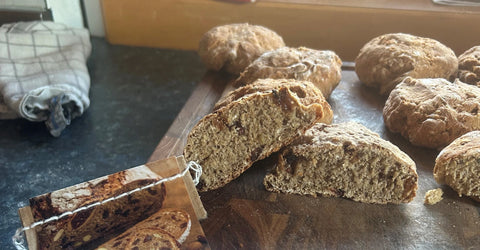 Aufgeschnittenes Früchtebrot mit sichtbaren Fruchtstücken auf Holzbrett, weitere Laibe und Leinentuch im Hintergrund
