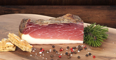 Angeschnittener Speck auf Holzbrett mit Zirbenzweig, Pfeffer, Salz und Schüttelbrot